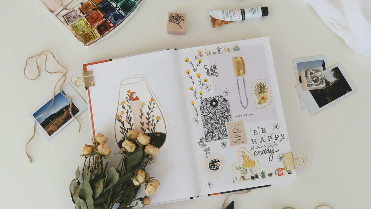 Open notebook with artistic illustrations and decorative elements surrounded by paint supplies, photographs, and stamps, on a white surface.