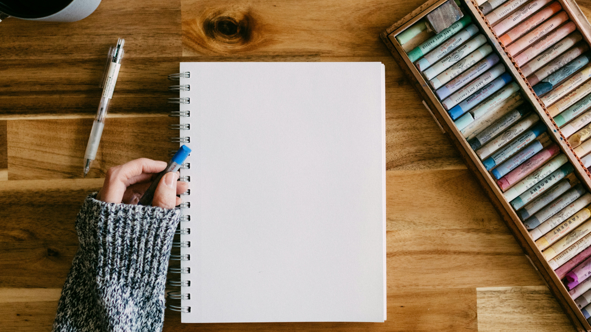 A top-down view of a person's hand holding a blue pastel next to a blank white spiral notebook on a wooden desk, with a box of colored pastels visible in the upper right.