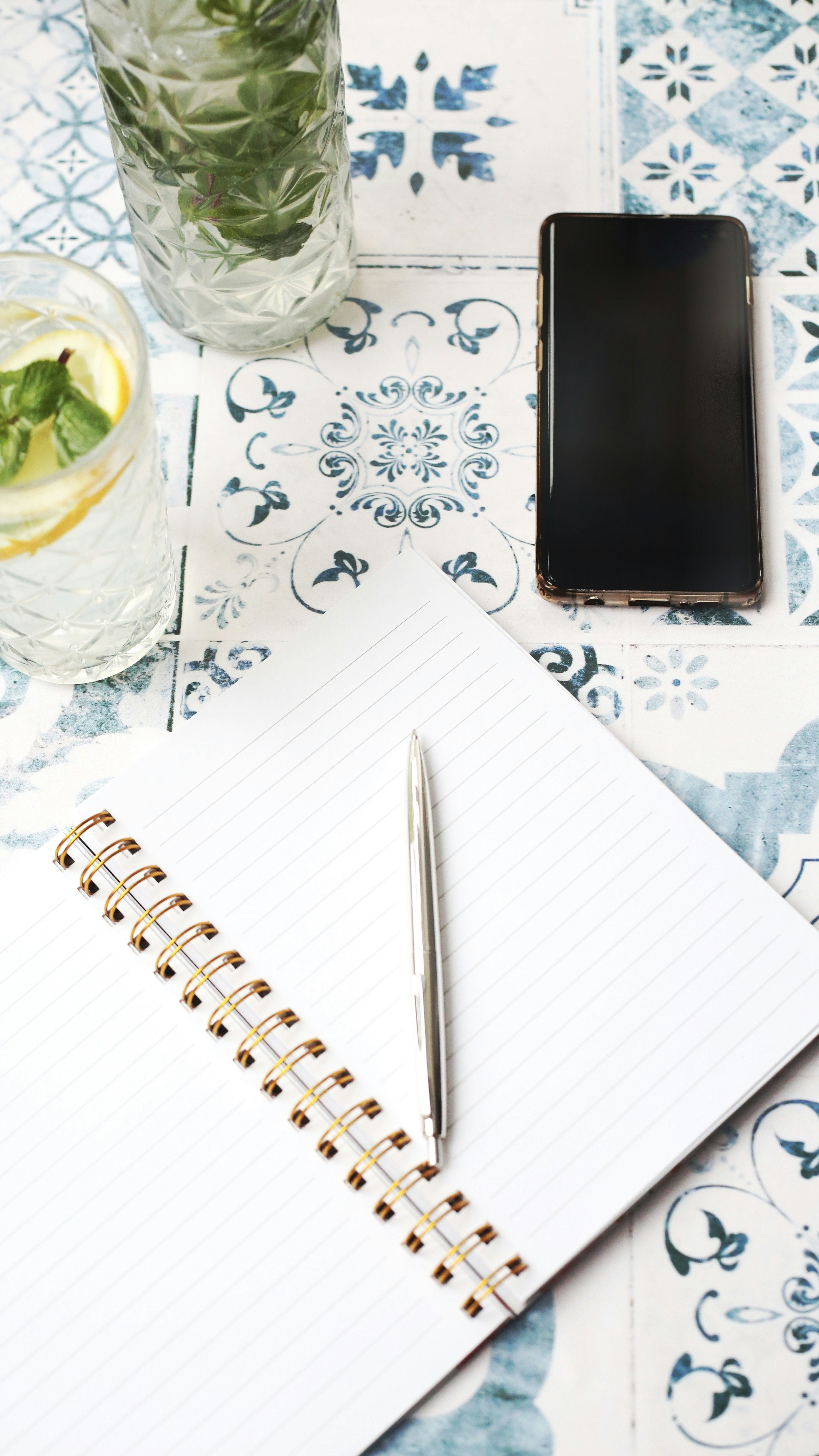 Tabletop scene with a notebook, pen, smartphone, and drink on a patterned tablecloth.